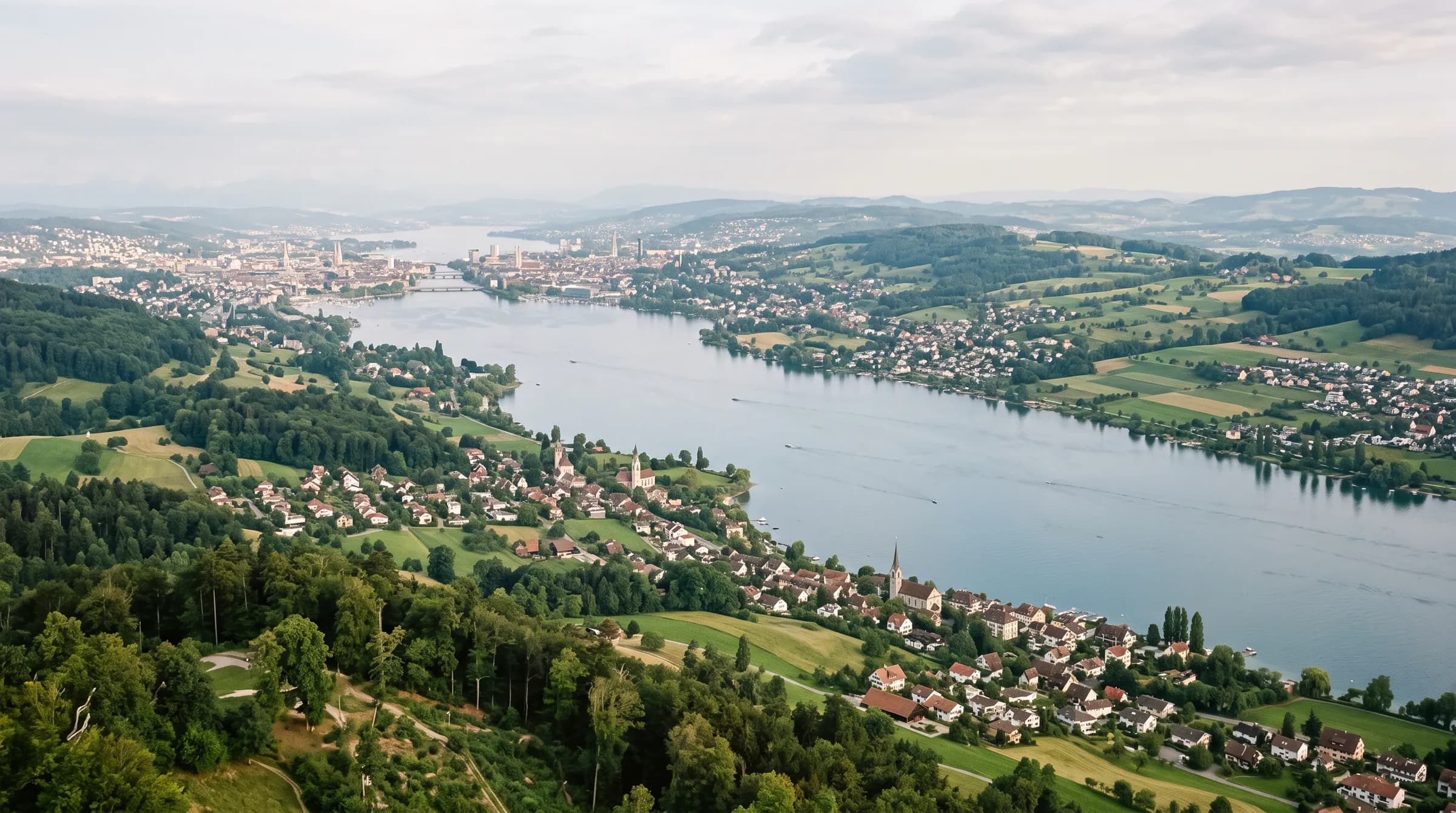 Blick über den Zürichsee auf Dörfer, Felder und bewaldete Hügel, im Hintergrund die Stadt.