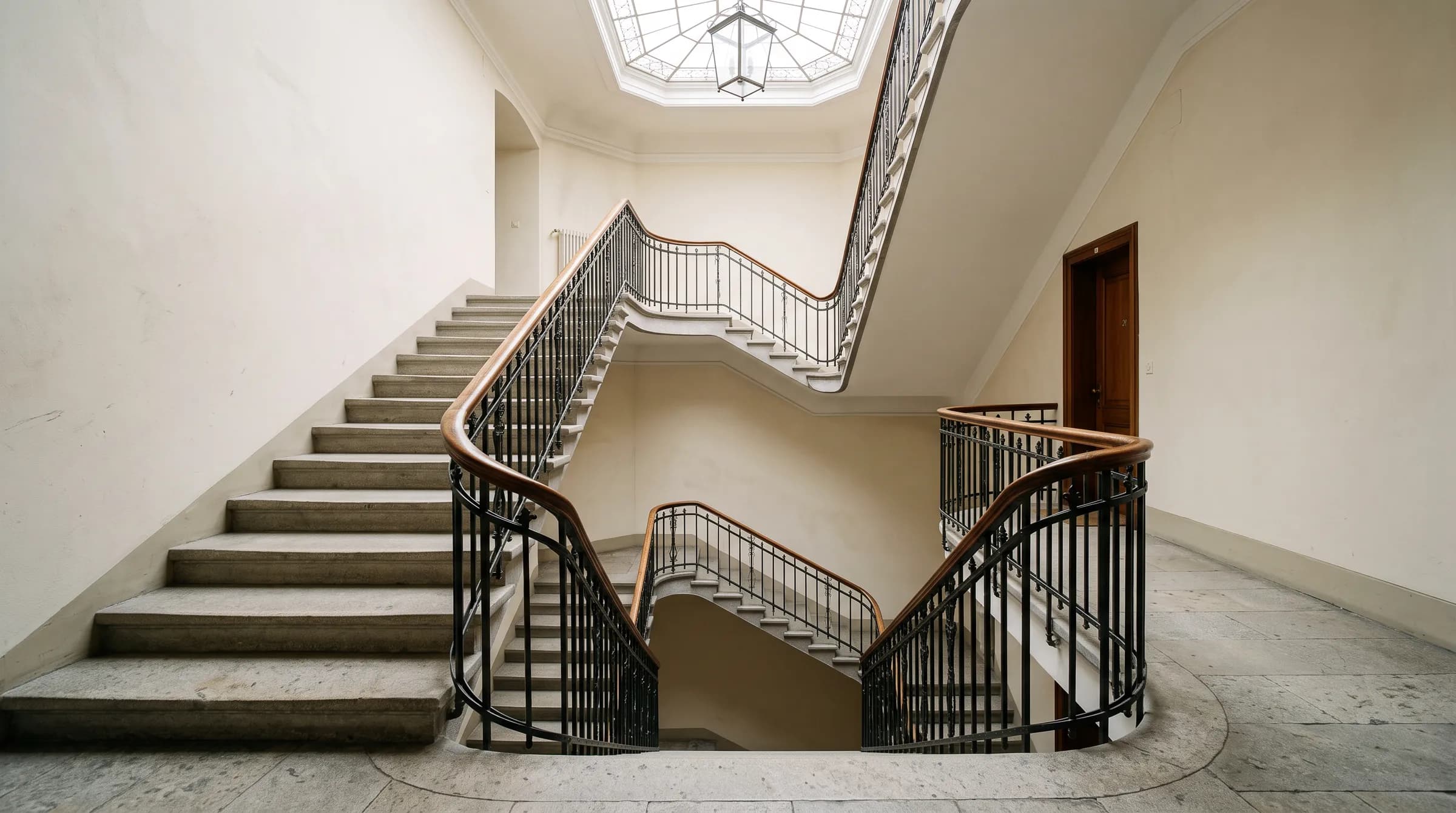 Treppenhaus eines Altbaus mit steinernen Stufen, schmiedeeisernem Geländer und Tageslicht aus einem Oberlicht.